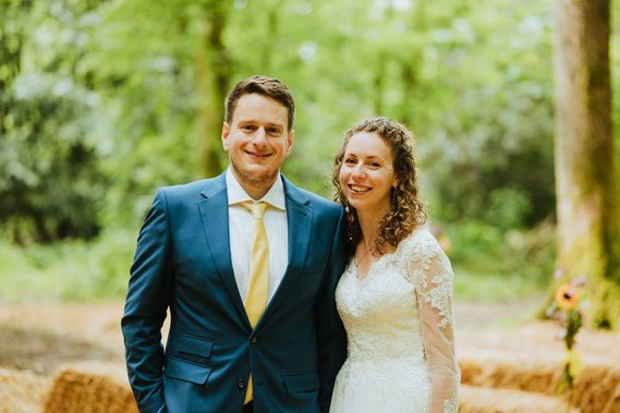 A couple stands together outdoors on their wedding day near a charming papermill in Kent. The man wears a blue suit with a yellow tie, and the woman wears a white lace dress. Trees and greenery are visible in the background.