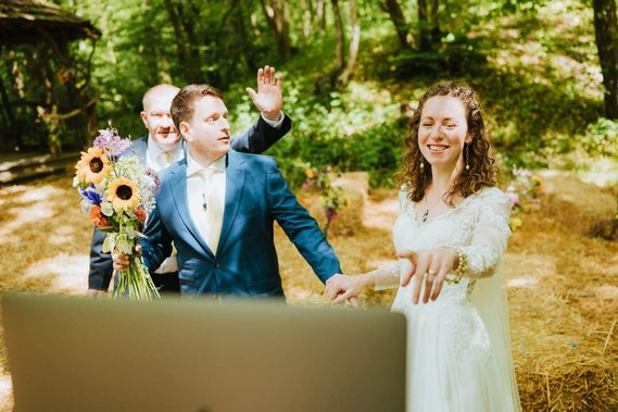 A bride and groom wave at a screen during an outdoor wedding ceremony in Kent. The groom holds a bouquet, and a third person stands nearby. The setting features trees and straw-covered ground, giving the scene a charming, pastoral feel reminiscent of an old papermill.