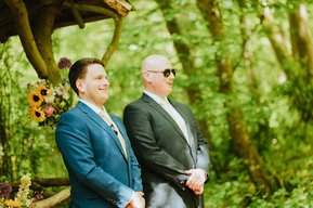 Two men in suits, one in blue and the other in black with sunglasses, stand outdoors in a forested area at a wedding. Sunflowers and greenery flourish around them, adding to the picturesque setting near a historic papermill in Kent.