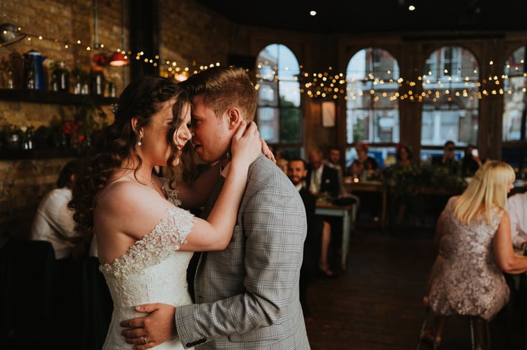 A couple shares a close dance at their wedding in a warmly lit venue with hanging string lights, brick walls, and other guests seated in the background. This romantic setting, reminiscent of a cozy spot tucked away in London, creates an unforgettable ambiance for their special day.