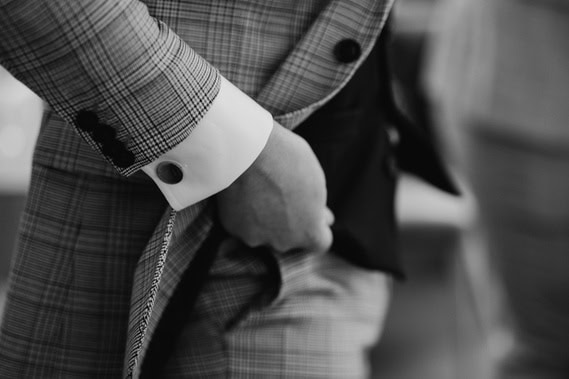 Close-up of a person in London adjusting a plaid suit jacket with a white shirt cuff and cufflink visible. The image, reminiscent of wedding elegance, is in black and white.
