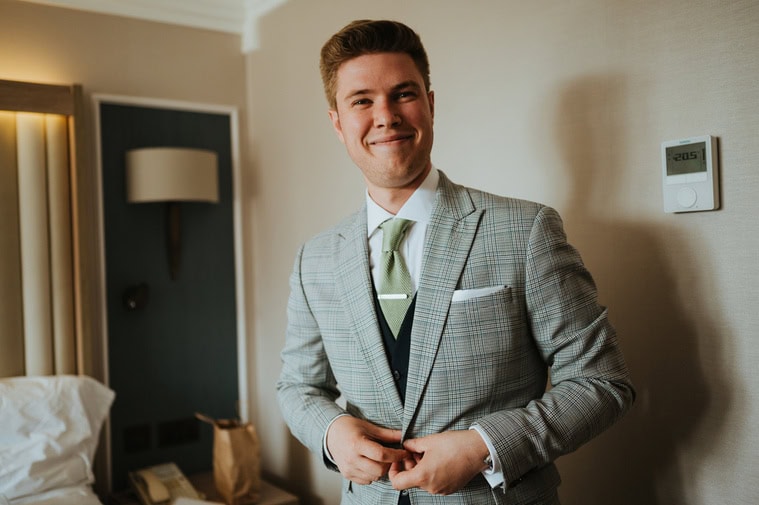 A man in a light gray checked suit and green tie smiles while standing in a room next to a bed and a wall-mounted thermostat, perhaps getting ready for a wedding in London.