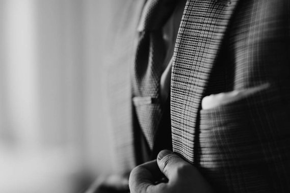 Close-up black and white image of a person adjusting their suit jacket, with one hand holding the lapel. The person is wearing a checked-pattern suit and a tie, preparing for what looks like an elegant London wedding.