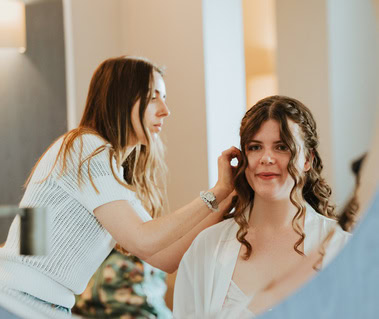 A hairstylist in a white top adjusts a woman's curly hair in front of a mirror, preparing her for her wedding day. The woman, draped in a white robe, radiates excitement as she gets ready for her special moment in London.