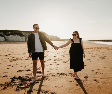 Two people holding hands walk barefoot on a sandy beach at Botany Bay, with a cliff in the background and the sun shining above—capturing a perfect moment of engagement through photography.