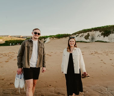 Two people standing on a sandy beach in Thanet, one holding shoes and the other holding sandals, with the stunning cliffs and lush greenery of Botany Bay in the background—a perfect moment captured through photography.