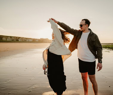 A man wearing sunglasses twirls a woman in a light jacket on Botany Bay beach during sunset, capturing a perfect moment for photography.