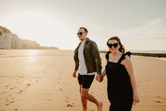 A man and a woman wearing sunglasses are walking hand in hand on a sandy beach with cliffs in the background on a sunny day, enjoying their engagement at Botany Bay.