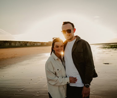 A couple stands close together on Botany Bay beach in Thanet at sunset, with the sun creating a halo-like effect above them. Wearing casual clothing and sunglasses, they look into each other's eyes, capturing the magic of their engagement.
