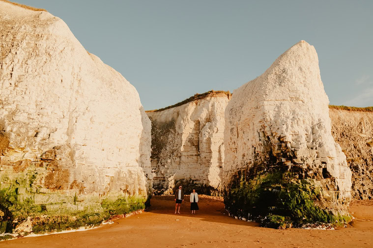 Two people stand on the sandy shore between large chalk cliffs under a clear sky, perfect for an engagement photoshoot at Botany Bay.
