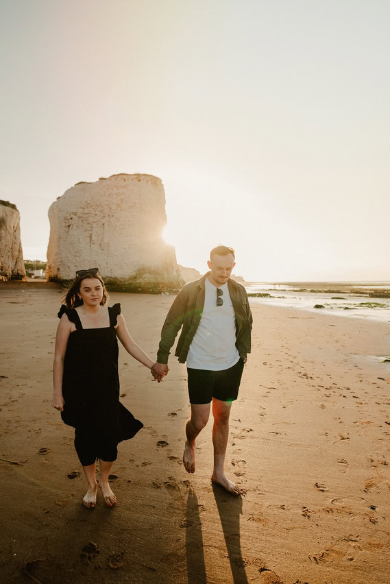 A couple holds hands while walking barefoot on the sandy shores of Botany Bay at sunset, with large rock formations of Thanet in the background, capturing a perfect engagement moment.