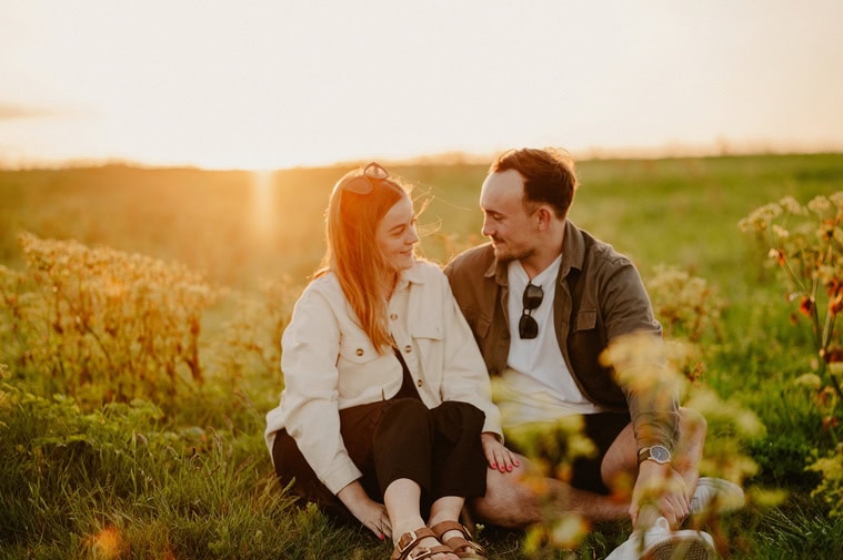 Two people sit on the grass in a field at sunset, smiling at each other. The sun casts a warm light, making everything look like a scene from an engagement photo shoot. They appear relaxed and content as if captured by a keen photography enthusiast.