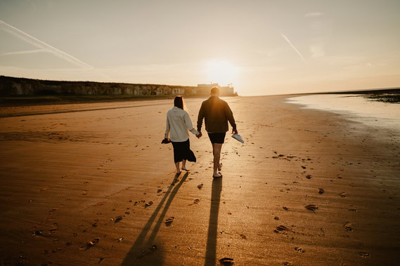 Two people walk hand-in-hand along a sandy beach at sunset in Thanet, leaving footprints behind them—a perfect moment captured by engagement photography.
