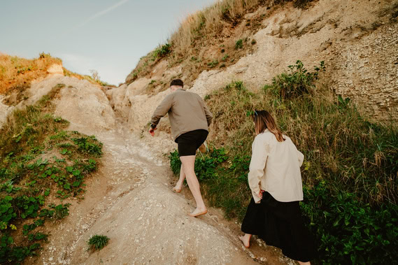 Two people, one barefoot and one in sandals, climb up a rocky hillside with sparse vegetation, perhaps imagining they're explorers at Thanet.
