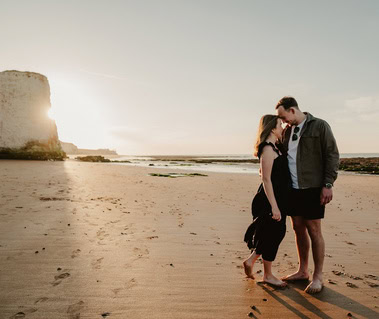 A couple stands closely together on a sandy beach at sunset, celebrating their engagement with the stunning cliffs and ocean of Botany Bay, Thanet in the background.