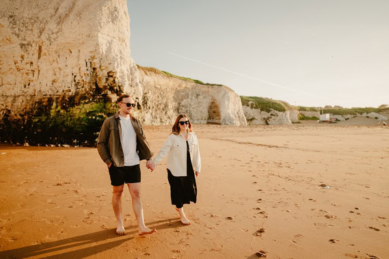 A couple is walking barefoot on a sandy beach near the stunning white cliffs of Thanet, holding hands and wearing sunglasses. The man is in shorts and a jacket, while the woman is in a black dress and light-colored jacket, creating a perfect scene for engagement photography.