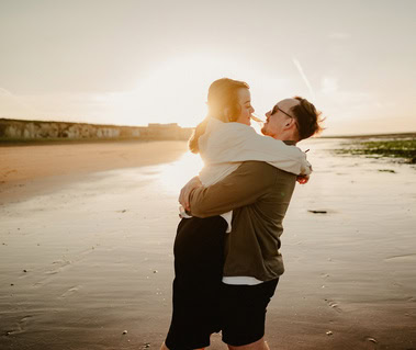 At Botany Bay in Thanet, a couple enjoys a moment during sunset. The man lifts and hugs the woman while both smile joyfully, celebrating their engagement by the serene shoreline.