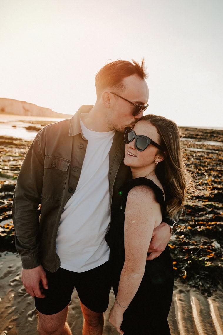 A man, wearing sunglasses and a jacket, kisses a woman, also wearing sunglasses, on the forehead while they stand on a rocky beach at sunset in Thanet, their engagement moment captured in stunning photography.