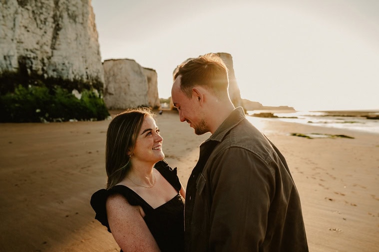 A couple stands close together on Botany Bay beach at sunset, facing each other and smiling, with large rock formations visible in the background. This stunning Thanet photography captures their joyful moment.