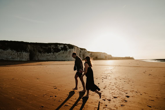 Two people walk barefoot along a sandy beach at sunset, casting long shadows. Cliffs of Botany Bay are visible in the background, perfect for stunning engagement photography.