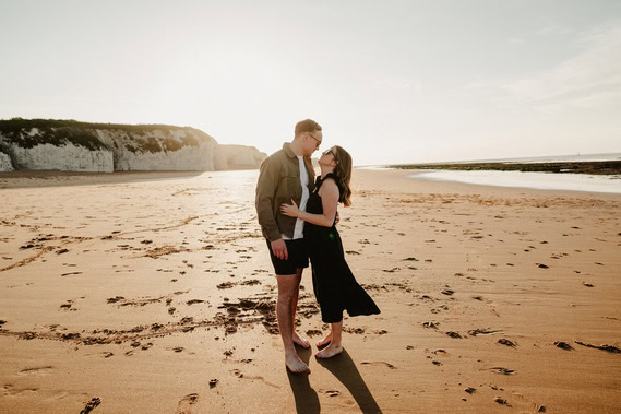 A couple is standing on a beach in Thanet, kissing with cliffs and the ocean in the background. The woman, wearing a black dress, embraces the man in a jacket and shorts. This tender moment captures their engagement perfectly through the lens of skilled photography.