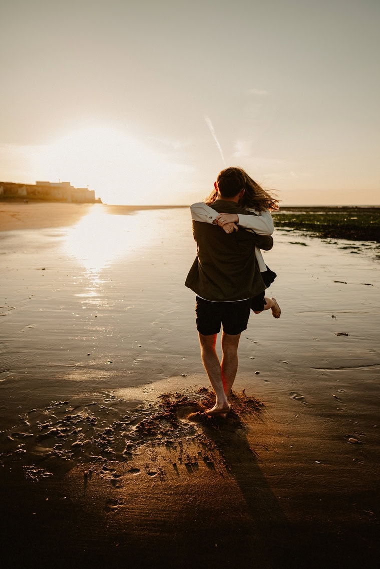A couple celebrating their engagement stands on a beach at sunset, holding each other in their arms as their shadows cast on the wet sand. The serene beauty of Botany Bay enhances the romantic atmosphere.