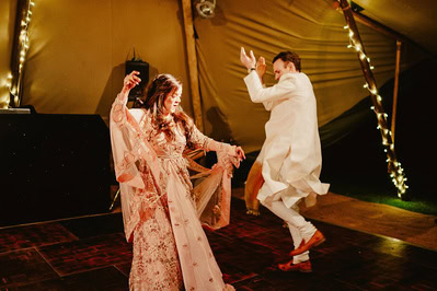 Two people dance energetically on a wooden floor under a tent adorned with string lights at a charming Kent wedding. They appear to be enjoying the moment, with one person wearing a light dress and the other in light-colored attire.