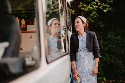 A woman in a blue dress and black jacket stands next to a vehicle, holding a phone and looking to the side, ready for the Kent wedding.