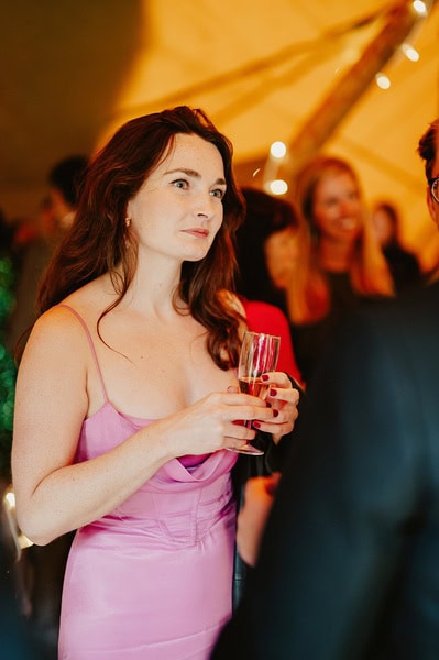 A woman in a pink dress holding a champagne flute stands in a tent at a Kent wedding, with other guests mingling in the background.