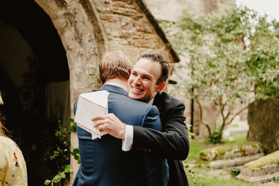 Two men dressed in suits hug warmly outside a stone building at a Kent wedding, one holding an envelope. The surrounding area includes greenery and another person partially visible on the left.