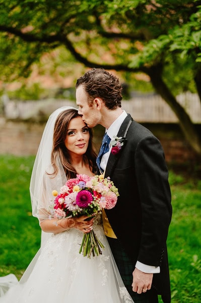 A couple on their Kent wedding day pose outdoors; the groom kisses the bride's forehead while she holds a bouquet of flowers.
