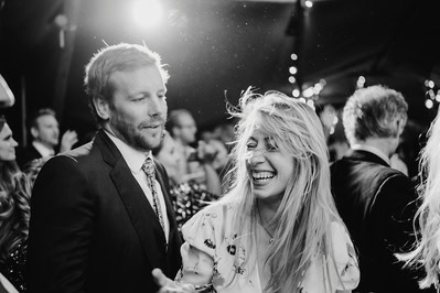 A black and white photo captures a man in a suit and a woman smiling at what appears to be a Kent wedding, with string lights illuminating the crowded event in the background.