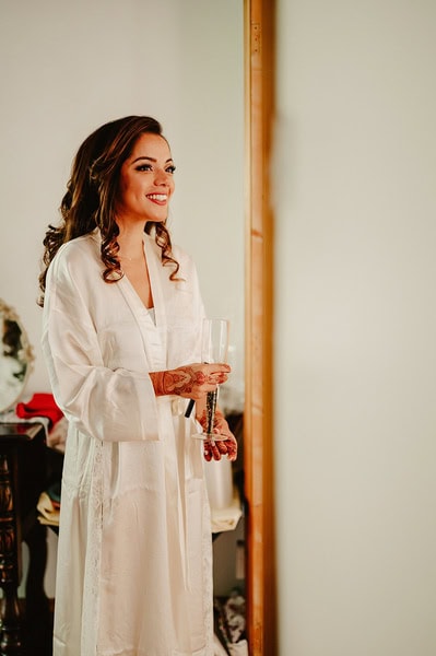 A woman in a white robe smiles while holding a glass of champagne, standing in a beautifully decorated room with a wooden door and various items on the table in the background, capturing the joy of her Kent wedding day.