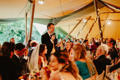 A man in a suit speaks into a microphone, addressing a group of seated guests under a tent with string lights. The event appears to be a formal gathering, possibly a Kent wedding reception.