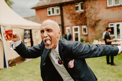 A man in a suit and bow tie poses playfully with his tongue out and holding a glass of drink, in front of a building during an outdoor Kent wedding.