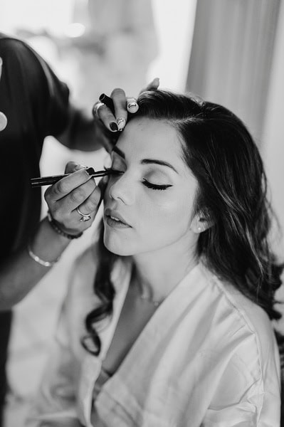 A woman with closed eyes is having makeup applied to her eyelids by another person, preparing for a beautiful Kent wedding.