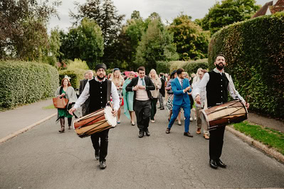 A group of people, including two drummers in traditional attire, walk down a residential street in Kent, with greenery and trees visible in the background, likely heading to a charming wedding ceremony nearby.