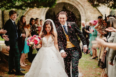 A bride and groom walk hand in hand while guests throw confetti at their Kent wedding. The bride holds a bouquet of flowers, and the groom wears a suit with a yellow vest. They are outside, with a stone archway behind them.