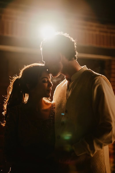 A man and woman stand close together, illuminated from behind, with the man kissing the woman's forehead. Both are dressed in traditional attire, capturing a tender moment at their Kent wedding.