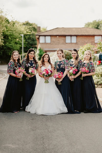 A bride in a white gown stands with five bridesmaids in matching navy dresses holding pink bouquets on a quiet Kent wedding street.
