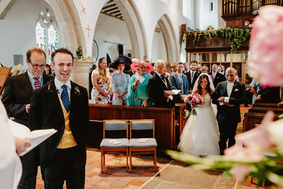 A bride walks down the aisle in a charming Kent church, accompanied by an older man. The groom and guests look on, some dressed in formal wear, aligned on pews.
