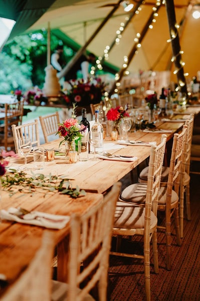 A long wooden table set for a Kent wedding, adorned with floral arrangements, glassware, wine bottles, and lit by string lights. Wooden chairs are neatly arranged on either side of the table.