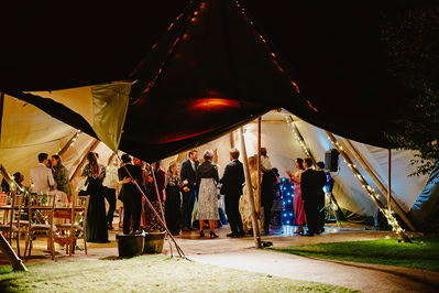 A group of people are gathered inside a large, illuminated tent at night, engaging in conversation and socializing during a lively Kent wedding.