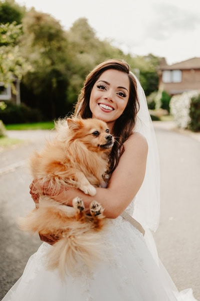 A woman in a wedding dress happily holding a small fluffy dog outdoors, surrounded by the greenery and charming houses typical of a Kent wedding.