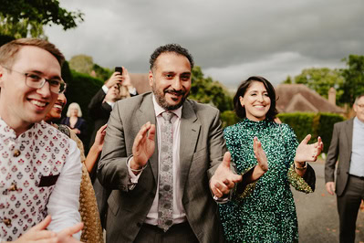 A joyous group of people, including a man in a suit and tie, are clapping and smiling outdoors at a charming Kent wedding, with trees and houses in the background.