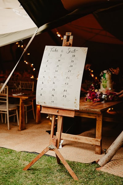 A seating chart on an easel stands outdoors under a tent at this charming Kent wedding, listing table numbers and guest assignments. Wooden tables and string lights are visible in the background, creating a warm, inviting atmosphere.
