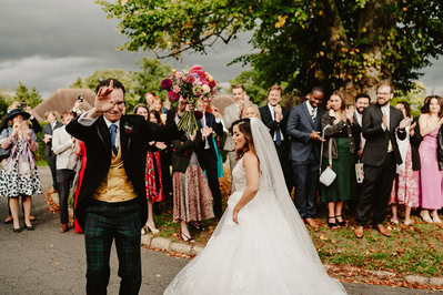A newlywed couple walks away from their Kent wedding ceremony smiling and waving. The bride holds a bouquet, and guests stand around clapping and taking photos. Trees and an overcast sky are in the background.