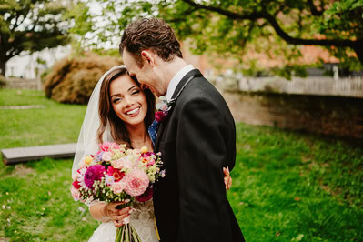 A bride holding a colorful bouquet smiles while being embraced by a groom in a suit outdoors. Both stand on a green lawn with trees and a brick wall in the background, capturing the joy of their Kent wedding.