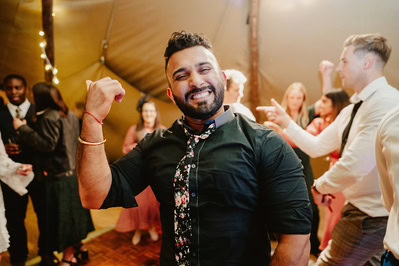 A man in a dark shirt and floral tie poses confidently with a flexed arm at a lively indoor Kent wedding. Other guests are seen laughing and socializing in the background.