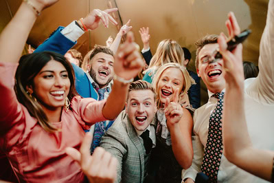 A group of people dressed in formal attire are joyfully celebrating a Kent wedding, with raised hands and wide smiles under a warmly lit setting.